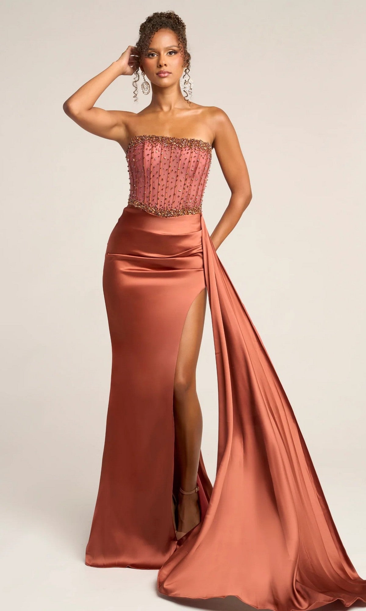 A woman poses against a plain background in the Portia and Scarlett XO040 long formal dress, featuring a bronze strapless beaded corset bodice, satin skirt with high slit, and draped sash.